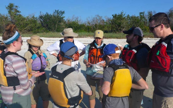A group of young people and their parents wear life jackets and stand in a circle on a beach.